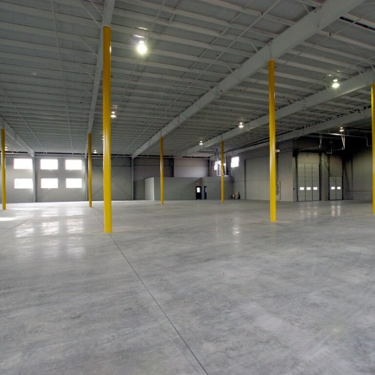 Interior of a vacant warehouse with 2 yellow support columns and light coming in through windows on the back wall.