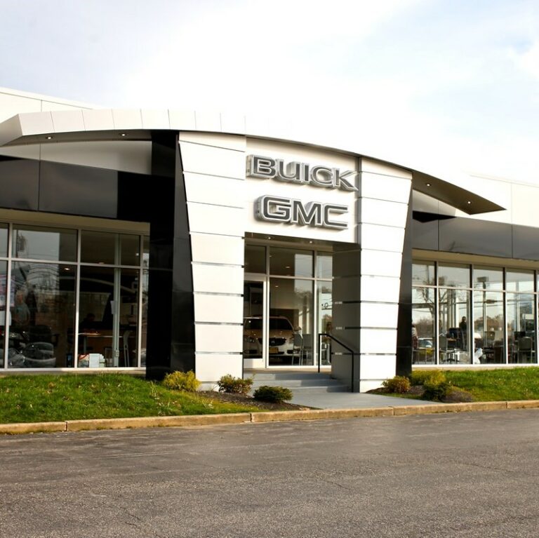 Front entrance to a Buick and GMC dealership on a partly cloudy day. Large glass windows on each side of the door.
